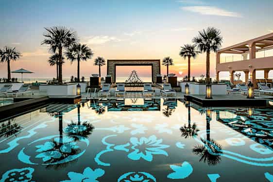 Bild av en pool med dekorativt mönster och omgivande solstolar, palmer och solnedgång i bakgrunden. (AI-genererad text)