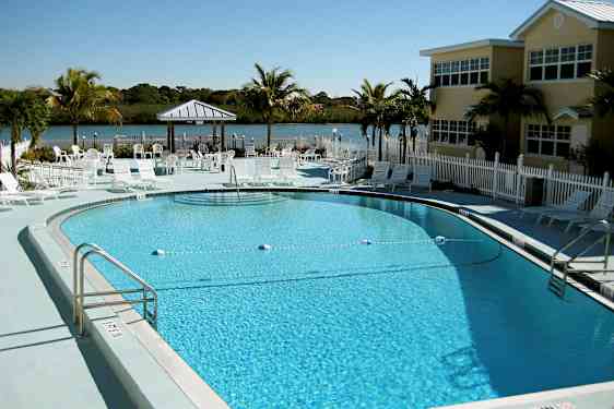 Bild av en utomhuspool omgiven av solstolar och parasoller. I bakgrunden syns en liten sjö omgiven av grönska. (AI-genererad text)