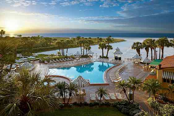 Bild av ett poolområde med solstolar och parasoll, omgiven av palmer och en strand i bakgrunden. (AI-genererad text)