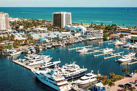 En småbåtshamn med lyxyachter och båtar bredvid urbana byggnader och grönområden, med havet i bakgrunden. (AI-genererad text)