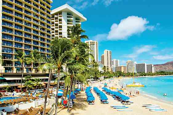 En sandstrand med solstolar och blå parasoll, omgiven av höga hotellbyggnader och palmer. (AI-genererad text)