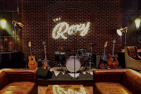 En scen med musikinstrument som trummor och gitarrer framför en tegelvägg med en lysande skylt som säger "The Roxy". (AI-genererad text)
