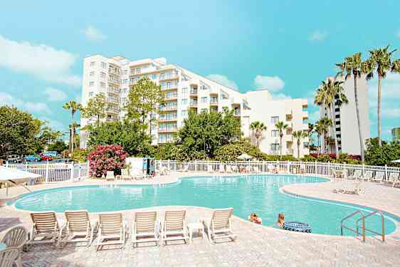 En utomhuspool framför en vit byggnad omgiven av träd och solsängar, med människor i vattnet och klarblå himmel ovanför. (AI-genererad text)