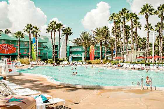 En utomhuspool omgiven av solstolar, parasoller och palmer. I bakgrunden syns en byggnad med färggranna detaljer. (AI-genererad text)