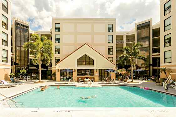 Bild av ett hotell med en pool i förgrunden. Personer badar och solar vid poolområdet. Hotellets fasad syns i bakgrunden med palmer i närheten. (AI-genererad text)