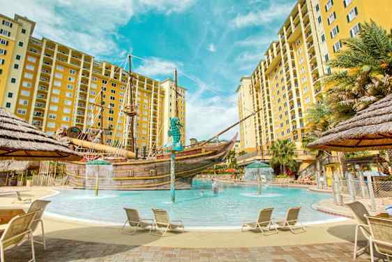 En pool omgiven av solstolar och gula flerbostadshus med ett piratskepp i mitten. (AI-genererad text)