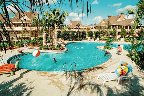 En utomhuspool omgiven av palmer och solstolar, med personer som badar och leker i vattnet. Byggnader syns i bakgrunden. (AI-genererad text)