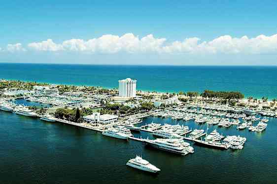 En marina med båtar och yachter vid en kust med en byggnad och ett hotell i bakgrunden. (AI-genererad text)