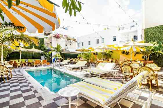 En ljus och färgglad utomhuspool med omgivande solstolar, parasoller och sittplatser omgiven av grönska och vita byggnader. (AI-genererad text)