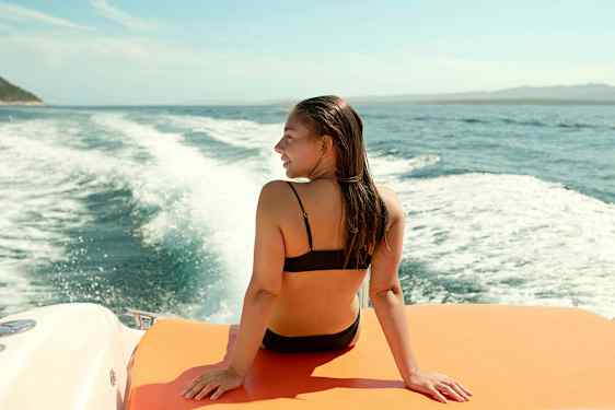 En person sitter på en orange yta på en båt med ryggen mot kameran, med havet och ett böljande spår i vattnet bakom. (AI-genererad text)