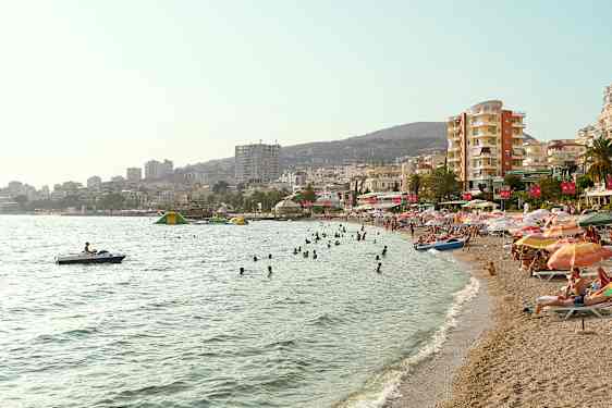 En strand med badande människor och parasoller, samt byggnader i bakgrunden. (AI-genererad text)