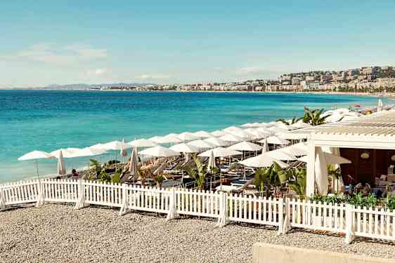 En solig strand med blått hav, parasoller och sittplatser omringade av ett vitt staket. (AI-genererad text)