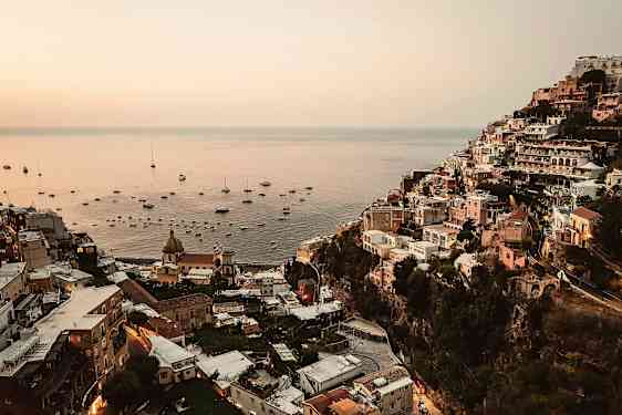 Bild av Positano under solnedgång med byggnader längs en sluttning och havet fullt av båtar. (AI-genererad text)