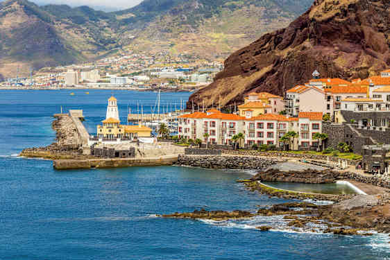 En kust med byggnader som har röda tak, en hamn och kullar i bakgrunden. (AI-genererad text)