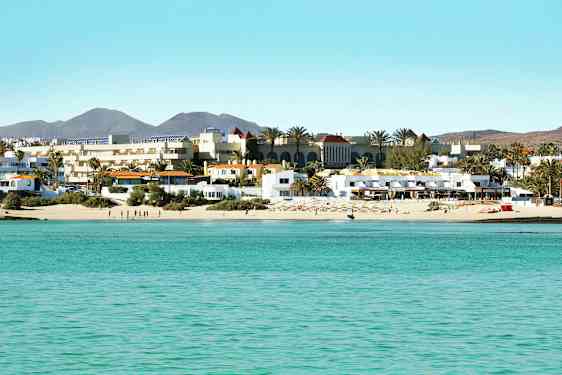 En strand med vit sand och turkost vatten framför en rad av byggnader och palmer. (AI-genererad text)