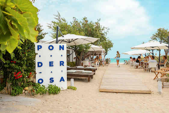 En restaurang vid stranden med bord och parasoller, en trägång leder mot vattnet. (AI-genererad text)