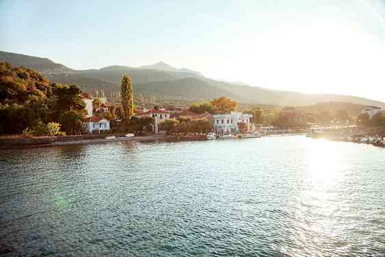 En liten by ligger vid en vattenkant med hus och träd omgivna av berg. Solen skiner över området. (AI-genererad text)