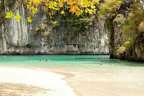Bilden visar en strand med klart turkost vatten omgiven av höga klippformationer täckta med växtlighet. Flera personer simmar i vattnet under en klar himmel. (AI-genererad text)