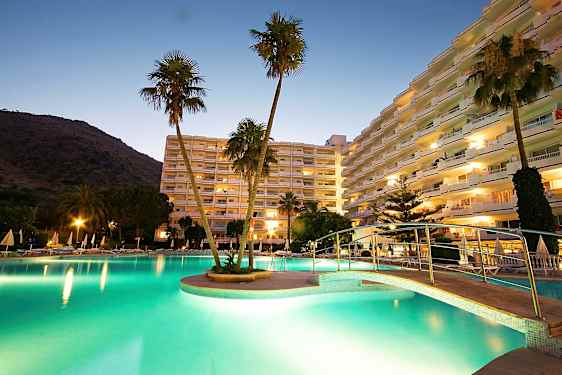 En bild på ett hotellområde vid skymning, med en upplyst pool, en bro och höga byggnader i bakgrunden. (AI-genererad text)