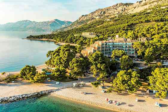 Flygbild över en strand med parasoller, byggnader omgivna av träd och berg i bakgrunden. (AI-genererad text)