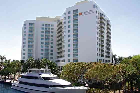 Bild av ett större vit byggnad med flera våningar och balkonger. Framför byggnaden syns en vit motorbåt förtöjd vid vattnet, samt träd och grönska. (AI-genererad text)