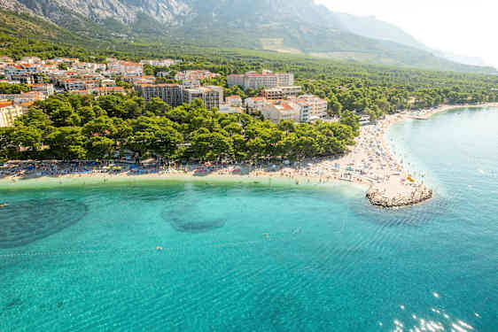 Luftbild av en kust trots klarblått vatten, strand med solstolar och en tät bebyggelse omgiven av skog och berg. (AI-genererad text)