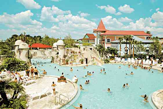 En utomhuspool omgiven av människor, solstolar och byggnader. (AI-genererad text)