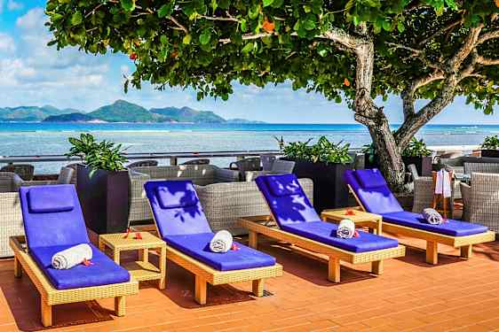 Solstolar med blå dynor och sammanrullade handdukar placerade vid ett hav med berg i bakgrunden. (AI-genererad text)