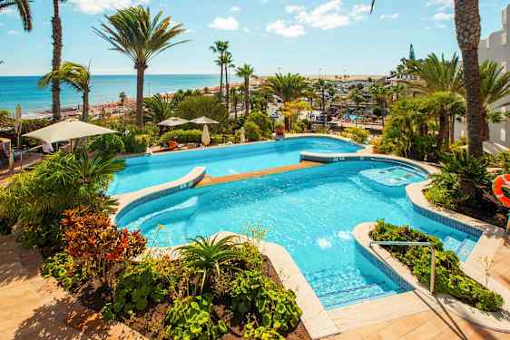 Utsikt över en pool omgiven av grönska och palmer, med havet i bakgrunden. (AI-genererad text)