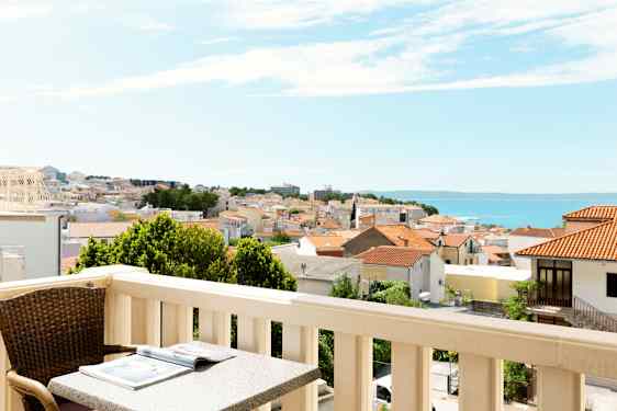 Balkong med bord och stol, utsikt över en stad med röda tak och havet i bakgrunden. (AI-genererad text)