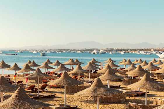 En strand med solstolar och parasoller av naturmaterial, med hav och båtar i bakgrunden. (AI-genererad text)