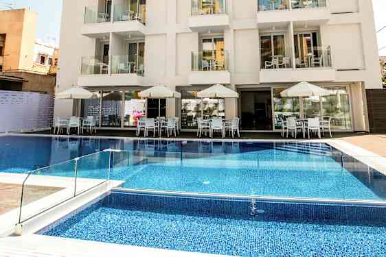 Ett hotell med flera våningar, balkonger och en pool framför byggnaden samt uteservering med bord och parasoller. (AI-genererad text)