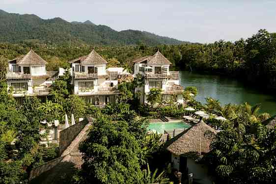 Ett hotellområde med hus i tropisk stil, omgiven av grönska och en flod i bakgrunden. (AI-genererad text)