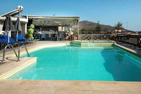 En utomhuspool med solstolar och parasoller intill samt en bar i bakgrunden. Ett berg syns i fjärran. (AI-genererad text)