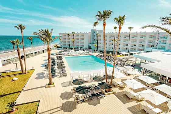 En vy över en resort med en stor pool, omgiven av solstolar och parasoller, med palmer och en byggnad i bakgrunden nära havet. (AI-genererad text)