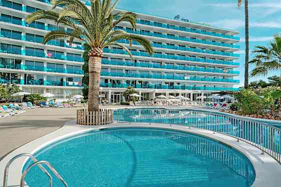 Bild av ett hotell med en pool, omgiven av solstolar och parasoller, med palmer och gröna växter. (AI-genererad text)