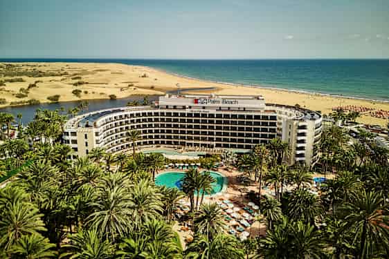 Palm Beach hotell med pool i förgrunden, omgiven av palmer, med sanddyner och hav i bakgrunden. (AI-genererad text)