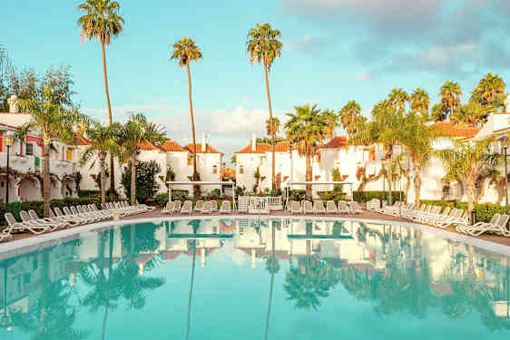 Hotellområde med vitputsade byggnader med röda tak, omgivna av palmer, och en stor pool med solstolar runt. (AI-genererad text)