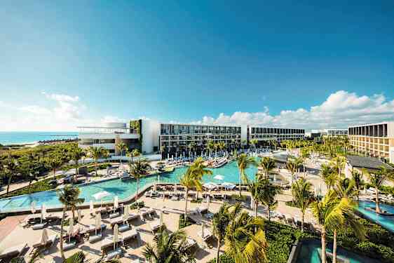 Ett modernt hotellkomplex med flera pooler, frodig vegetation och palmer, beläget vid kusten med utsikt över havet under en klarblå himmel. (AI-genererad text)