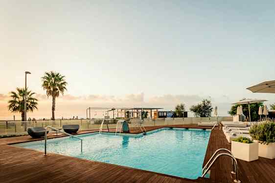 Bilden visar en utomhuspool med trädäck och solsängar vid kanten. I bakgrunden syns ett par palmer, en byggnad och havet, allt under en ljus kvällshimmel. (AI-genererad text)