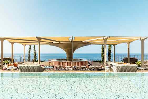 En pool med utsikt över havet och möblerade områden under skuggande tak. (AI-genererad text)