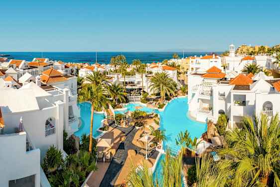 Poolområde med vita byggnader och röd tak, omgiven av palmer med utsikt över havet. (AI-genererad text)