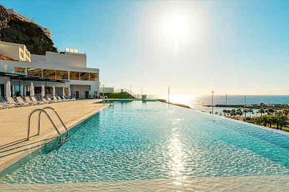 En infinitypool med spegelklart vatten, solsängar och parasoller på sidan, i ett hotellområde med utsikt mot havet och stranden. (AI-genererad text)