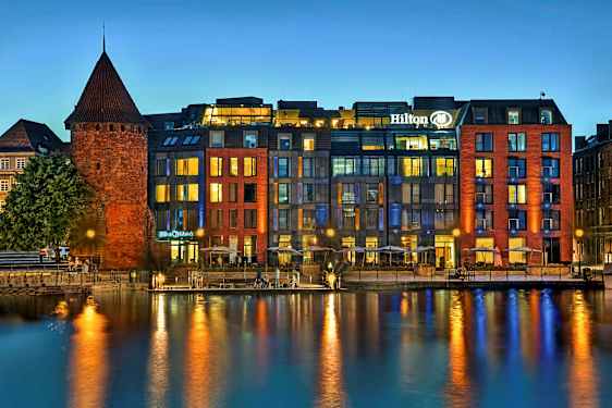 En historisk byggnad i tegel bredvid ett modernt hotell vid vattnet på kvällen. (AI-genererad text)
