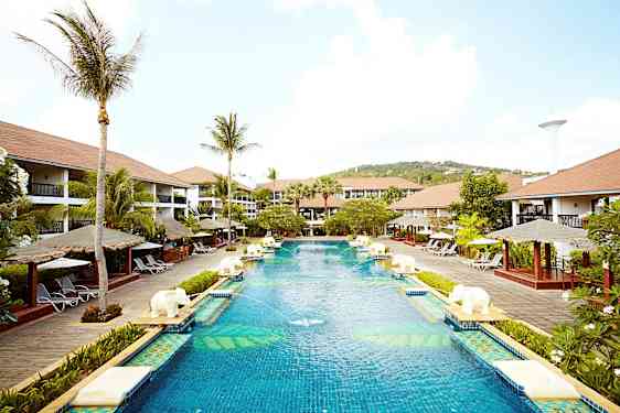 En stor pool på ett semesterortsområde omgiven av palmer, solstolar och bungalowliknande byggnader. (AI-genererad text)