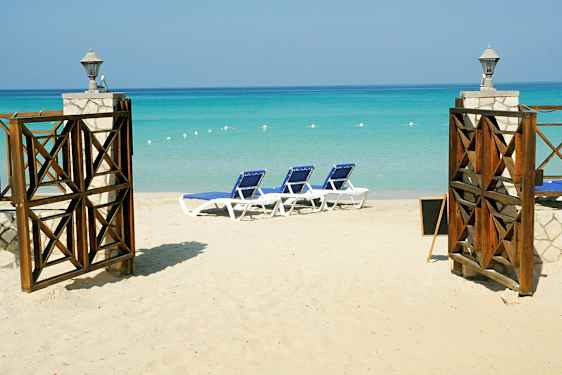 En sandstrand med solstolar och en öppnad port med utsikt över det turkosa havet. (AI-genererad text)