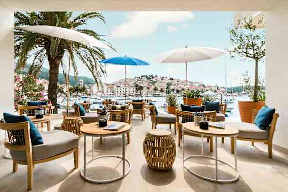 Uteservering med bord och stolar under parasoller, omgiven av palmer med utsikt över en pittoresk hamn och stad. (AI-genererad text)