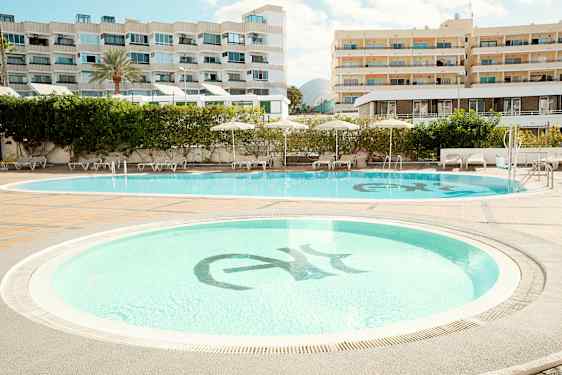 Två swimmingpooler omgivna av solstolar och parasoller, med hotellbyggnader i bakgrunden. (AI-genererad text)