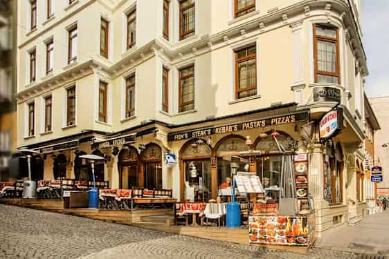 En gata med en byggnad som har restaurang med uteservering, skyltar för mat som kebab och pizza. (AI-genererad text)