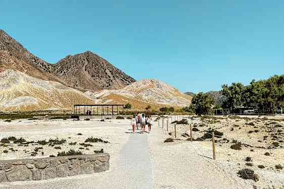 En torr landskapsvy med personer som går på en stig som leder mot några små byggnader. Berget syns i bakgrunden under en klarblå himmel. (AI-genererad text)